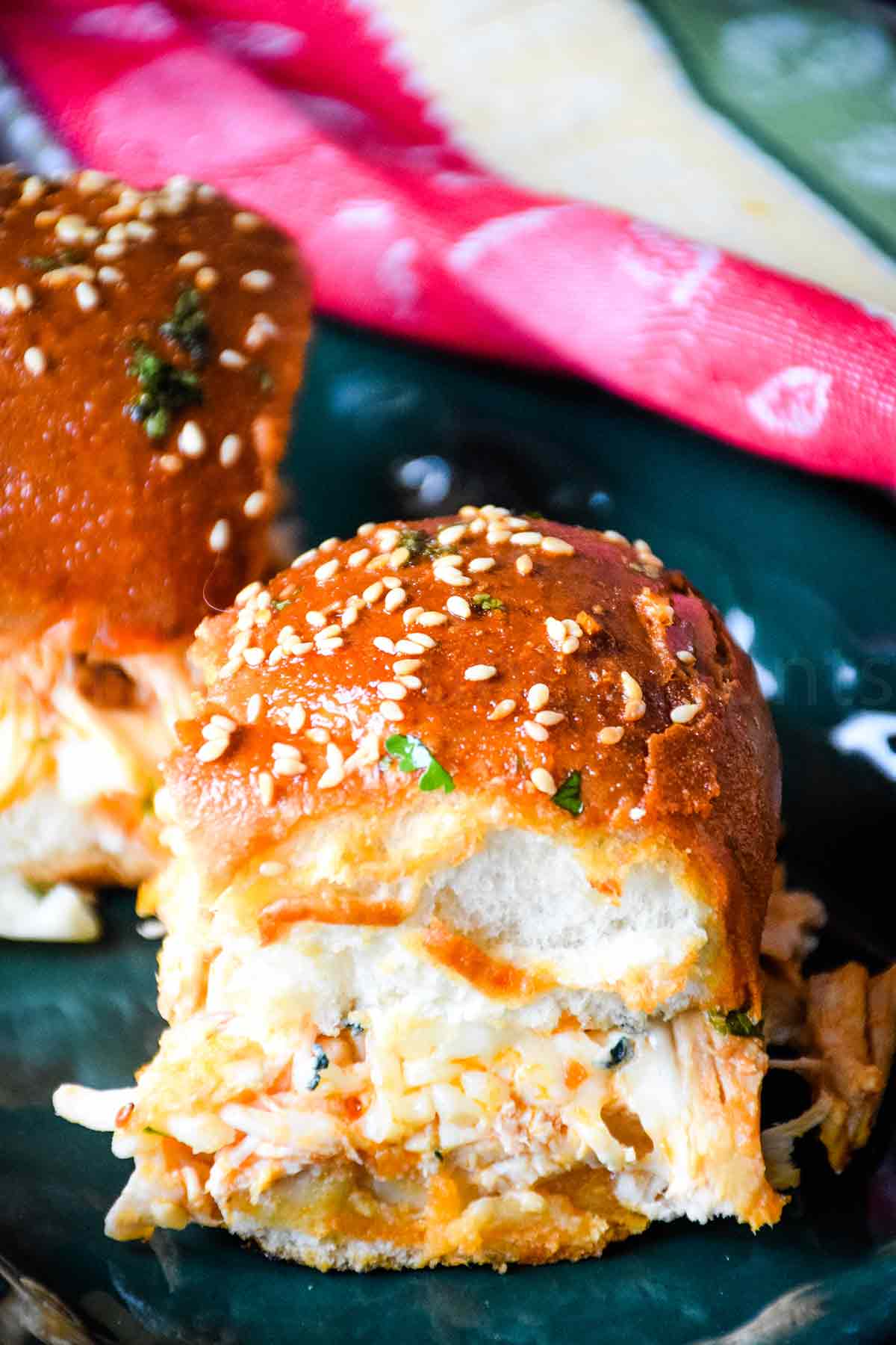 Close up of a buffalo chicken slider with a glossy, golden-brown bun topped with sesame seeds and chopped parsley, revealing shredded chicken and melted cheese inside.