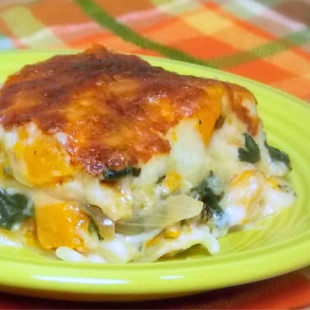 A slice of butternut squash lasagna served on a lime green plate, layered with golden roasted squash, spinach, onions, creamy cheese, and a browned cheesy topping.