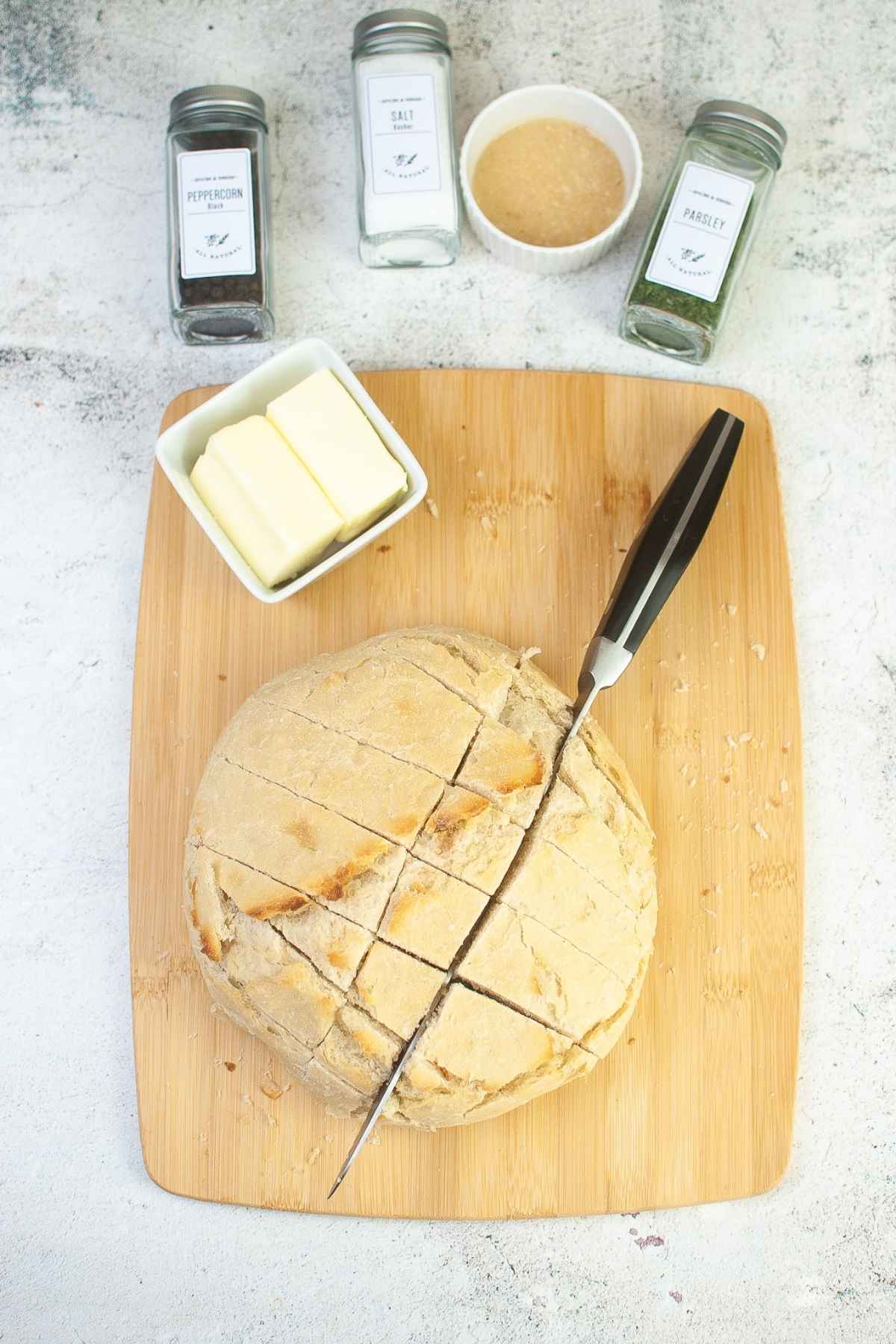Round loaf of bread being sliced in a crisscross pattern on a wooden cutting board with butter and seasonings nearby.
