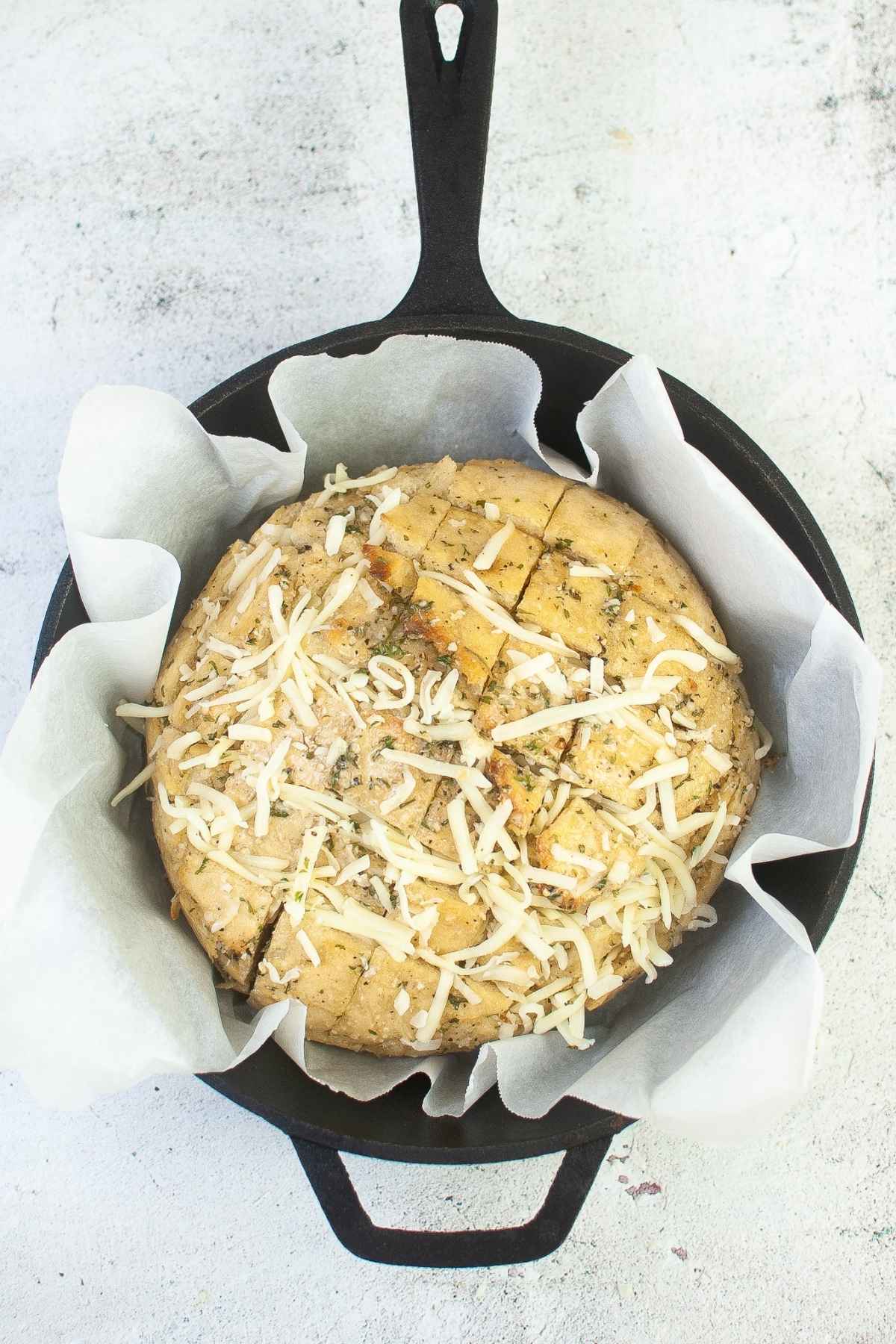 Overhead image of a round loaf of bread cut in a crisscross pattern in a parchment lined skillet sprinkled with mozzarella cheese and butter garlic herb mixture ready to be baked.