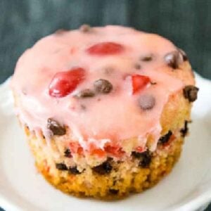 A close up of a cherry chocolate chip muffin topped with pink glaze, sitting on a white plate.