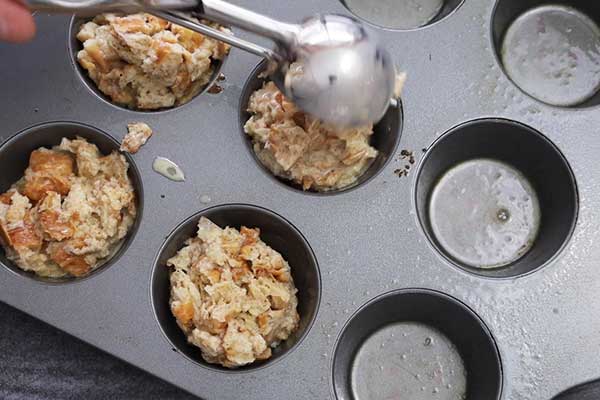 Muffin tin being filled with croissant muffin mixture using a large scoop.