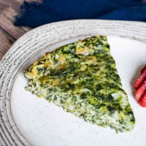 A slice of crustless spinach quiche on a plate with sliced strawberries on the side.