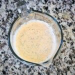 Glass measuring cup filled with creamy dill sauce, speckled with herbs, sitting on a gray and black speckled countertop.