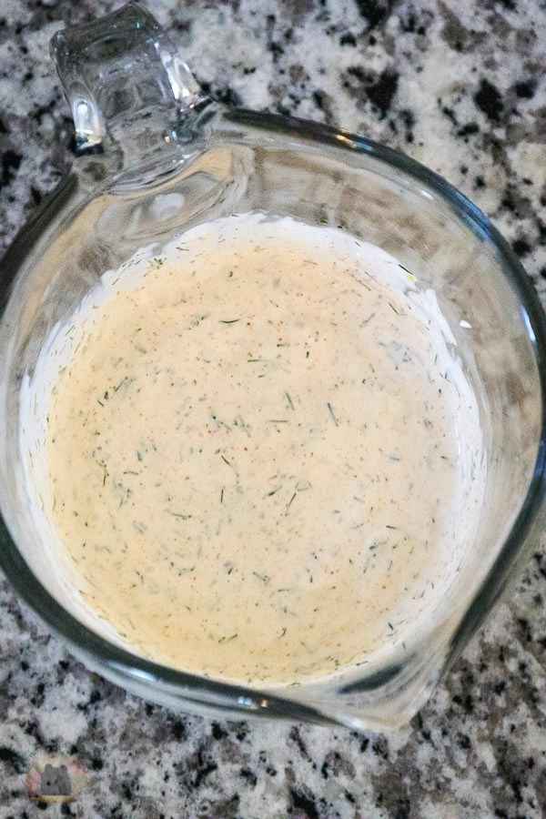 Glass measuring cup filled with creamy dill sauce, speckled with herbs, sitting on a gray and black speckled countertop.