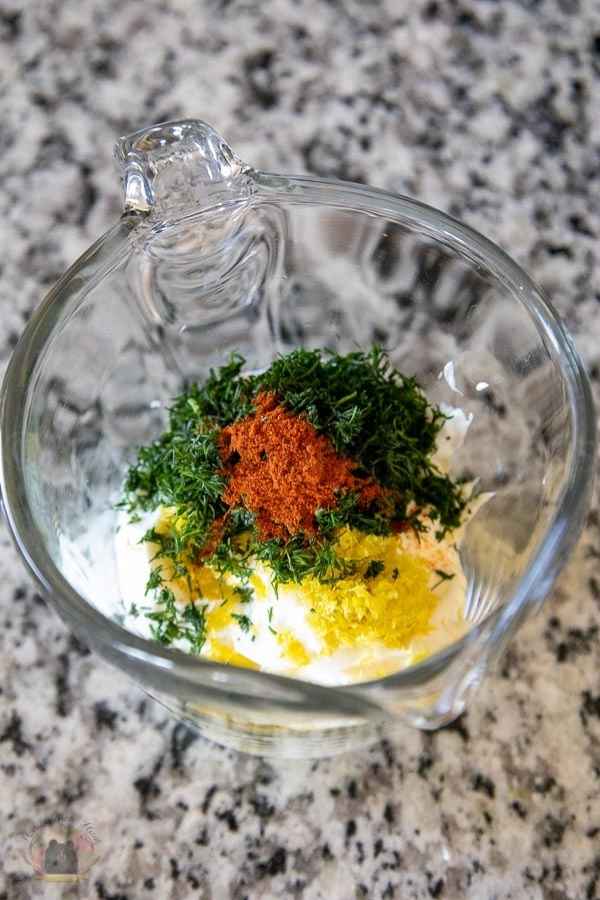 Unmixed sauce ingredients in a glass measuring cup, including fresh chopped dill, lemon zest, and smoked paprika, atop a creamy white base.