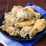 A black plate piled high with golden, crispy chicken wings coated in a generous layer of grated Parmesan cheese, set on a blue and white cloth napkin over a wooden table.