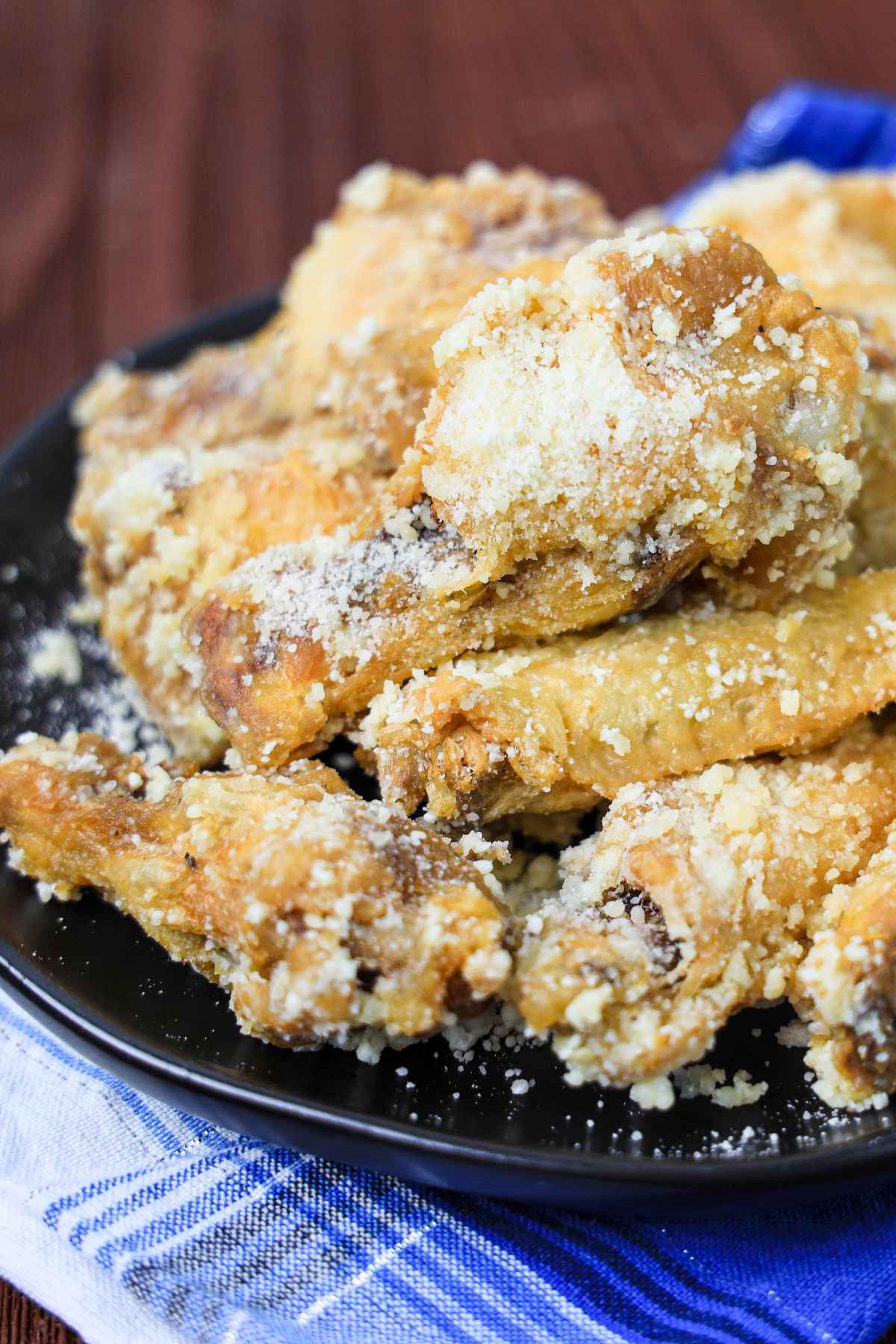 A black plate piled high with golden, crispy chicken wings coated in a generous layer of grated Parmesan cheese, set on a blue and white cloth napkin over a wooden table.