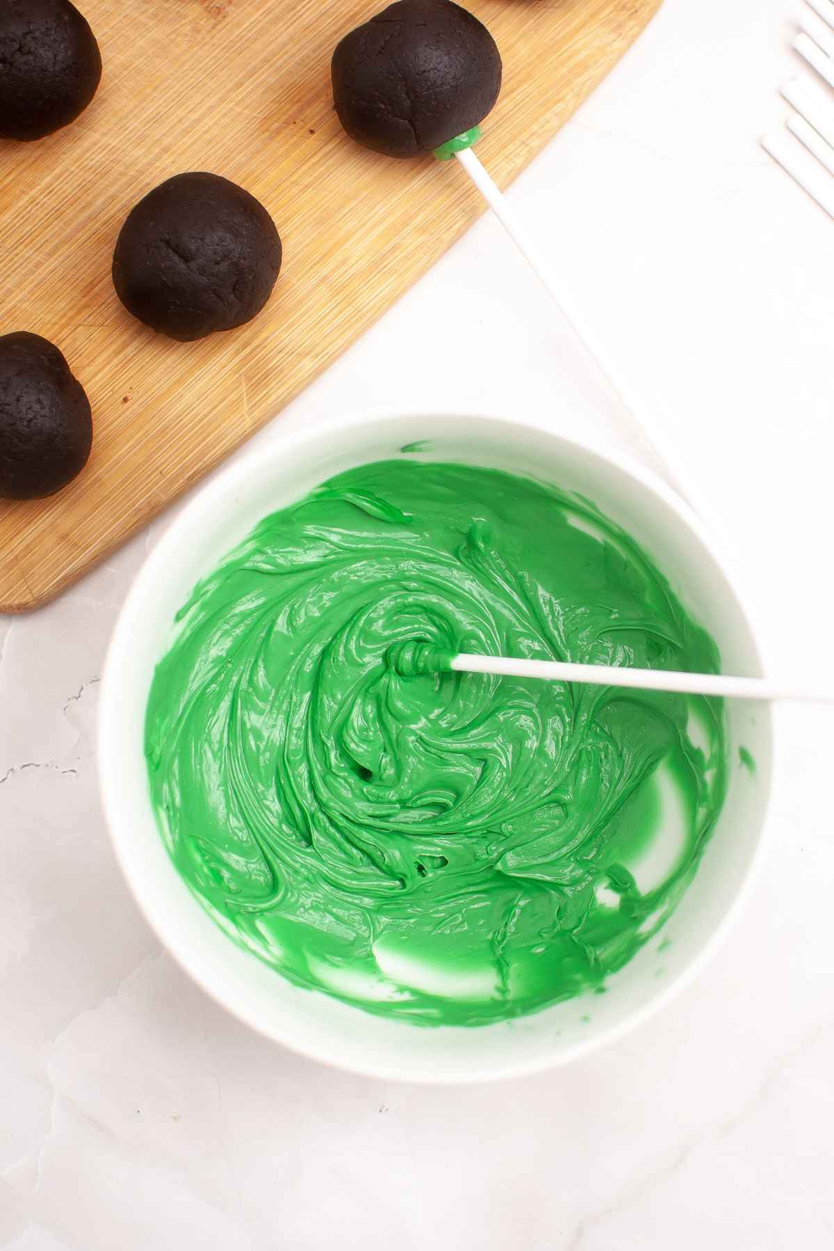 A bowl of melted green candy melts with a lollipop stick resting inside, surrounded by chocolate cake balls on a wooden board.
