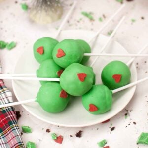 A plate of festive Grinch cake pops coated in green candy melts and topped with a red candy heart decoration.