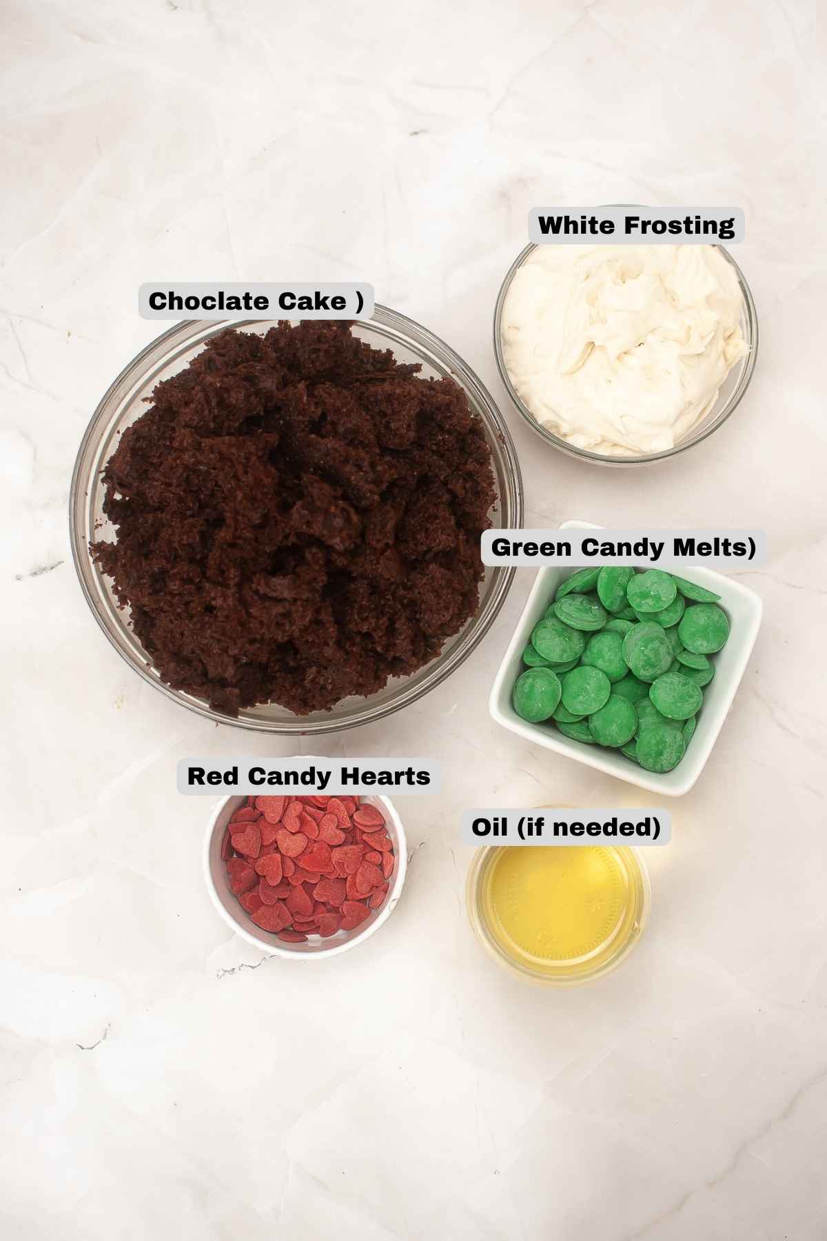 Overhead view of ingredients including crumbled chocolate cake, white frosting, green candy melts, red candy hearts, and oil in small bowls.