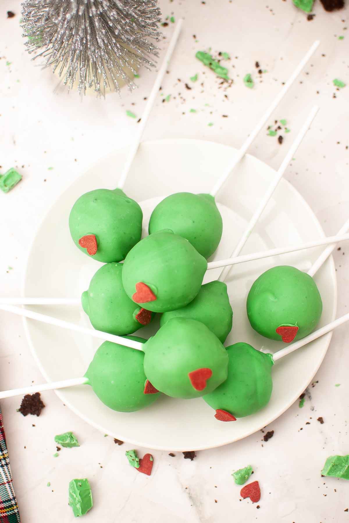 A plate of festive Grinch cake pops coated in green candy melts and topped with a red candy heart decoration.