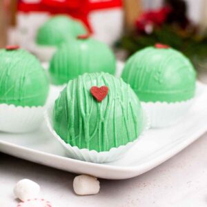 Festive green hot cocoa bombs decorated with red candy hearts, displayed on a white plate with holiday decor in the background.