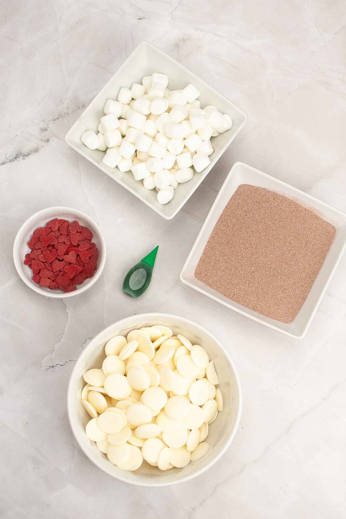 Overhead view of ingredients for Grinch hot cocoa bombs, including white chocolate wafers, mini marshmallows, hot cocoa mix, green food coloring and red heart sprinkles.