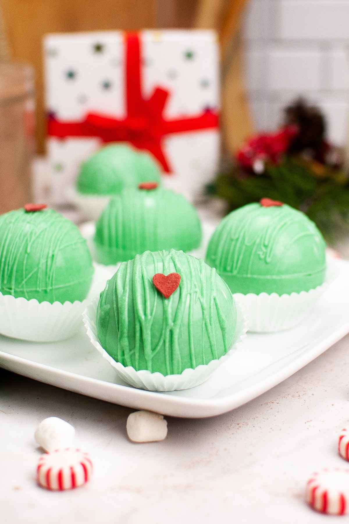 Festive green Grinch hot cocoa bombs decorated with red candy hearts, displayed on a white plate with holiday decor in the background.