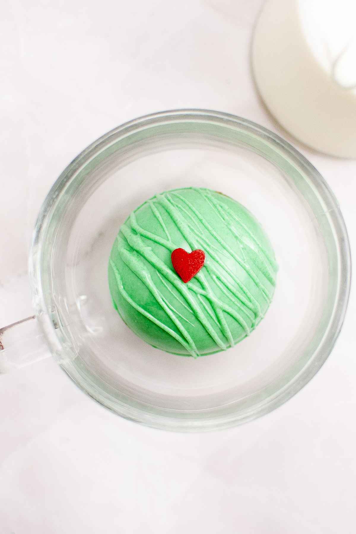 A green Grinch Hot Cocoa Bomb with a red heart on top sitting inside a clear mug, ready for hot milk to be poured over.