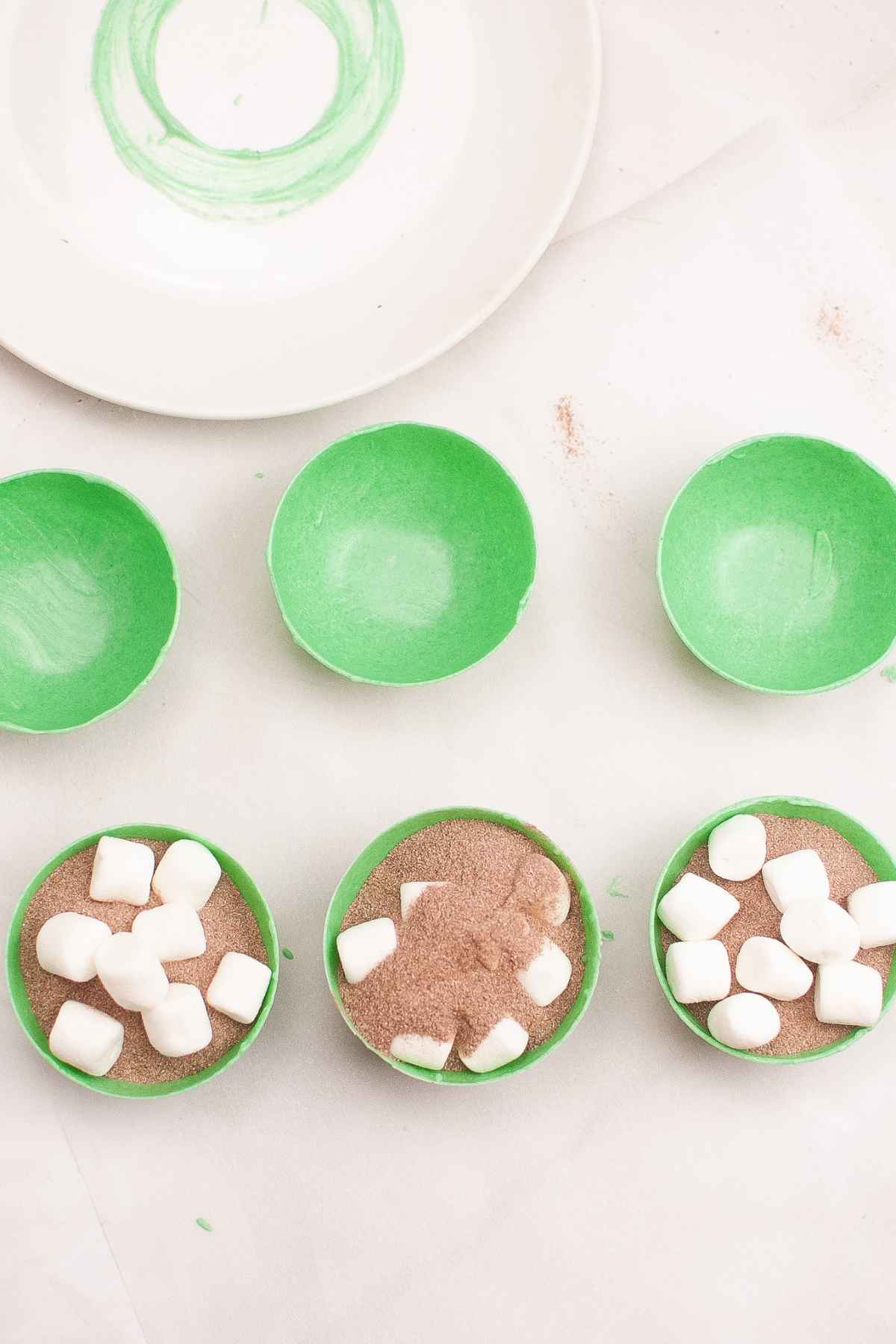 Green chocolate shells filled with hot cocoa mix and mini marshmallows, ready to be sealed to make hot cocoa bombs.