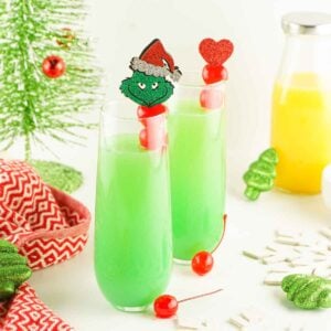 Festive green Grinch Mimosa in champagne flutes, garnishied with red cherries and holiday picks, surrounded by Christmas decorations and a bottle of orange juice.