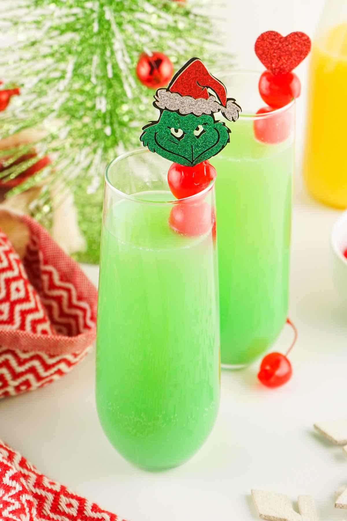 Festive green Grinch Mimosa in champagne flutes, garnishied with red cherries and holiday picks, surrounded by Christmas decorations and a bottle of orange juice.