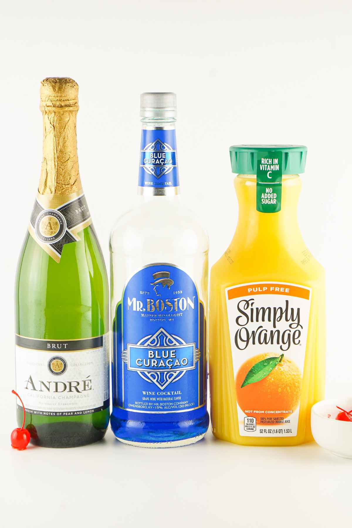 A bottle of champagne, blue curacao, and orange juice displayed side by side with maraschino cherries.