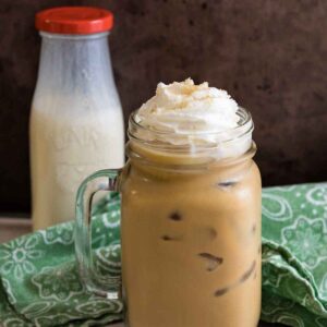 Glass mug of iced coffee topped with whipped cream and brown sugar garnish, with a milk bottle filled with almond coffee creamer in the background.