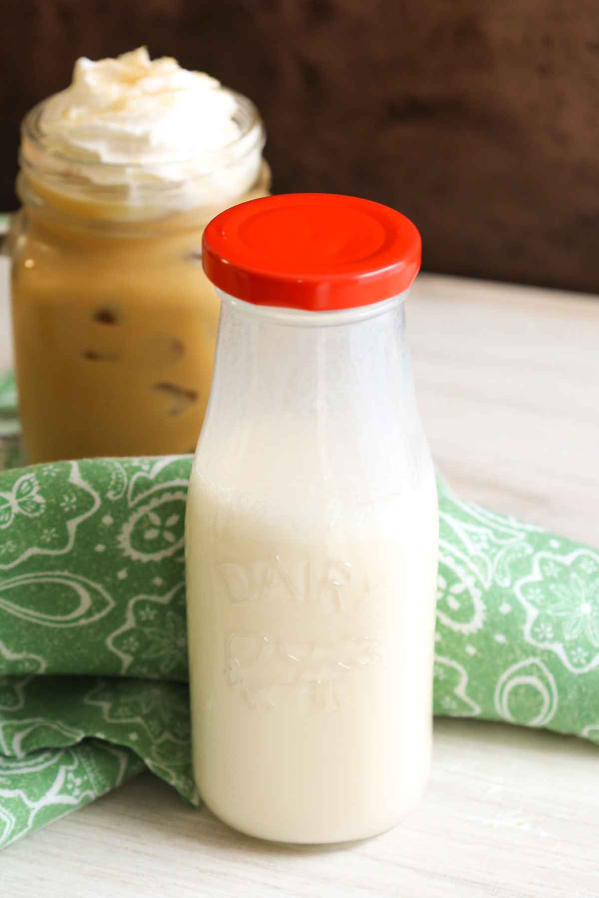 Close up of a glass milk bottle with a red cap, with a mug of iced coffee blurred in the background.