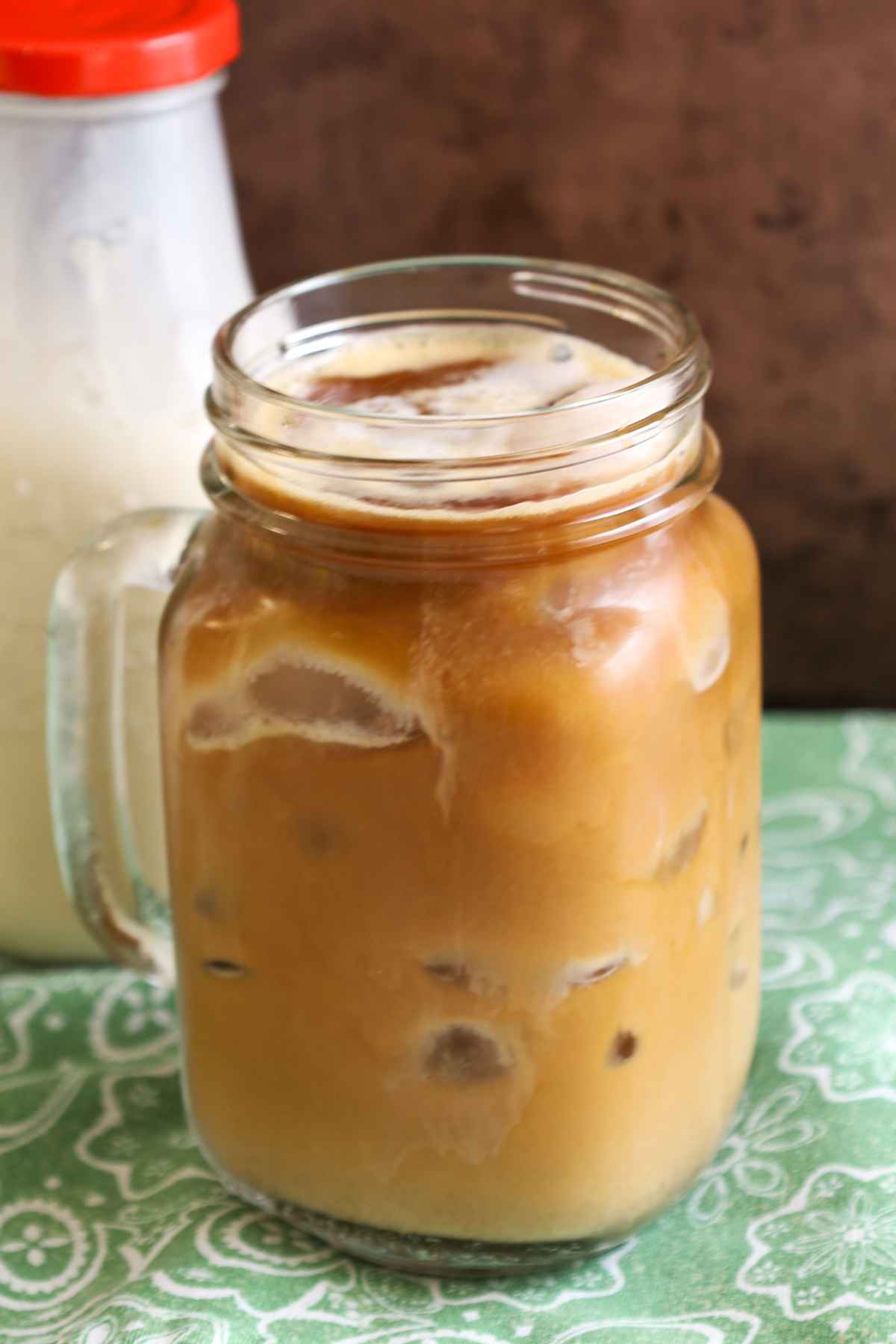 Glass muf of iced coffee with cream swirled in, sitting on a green patterned cloth.