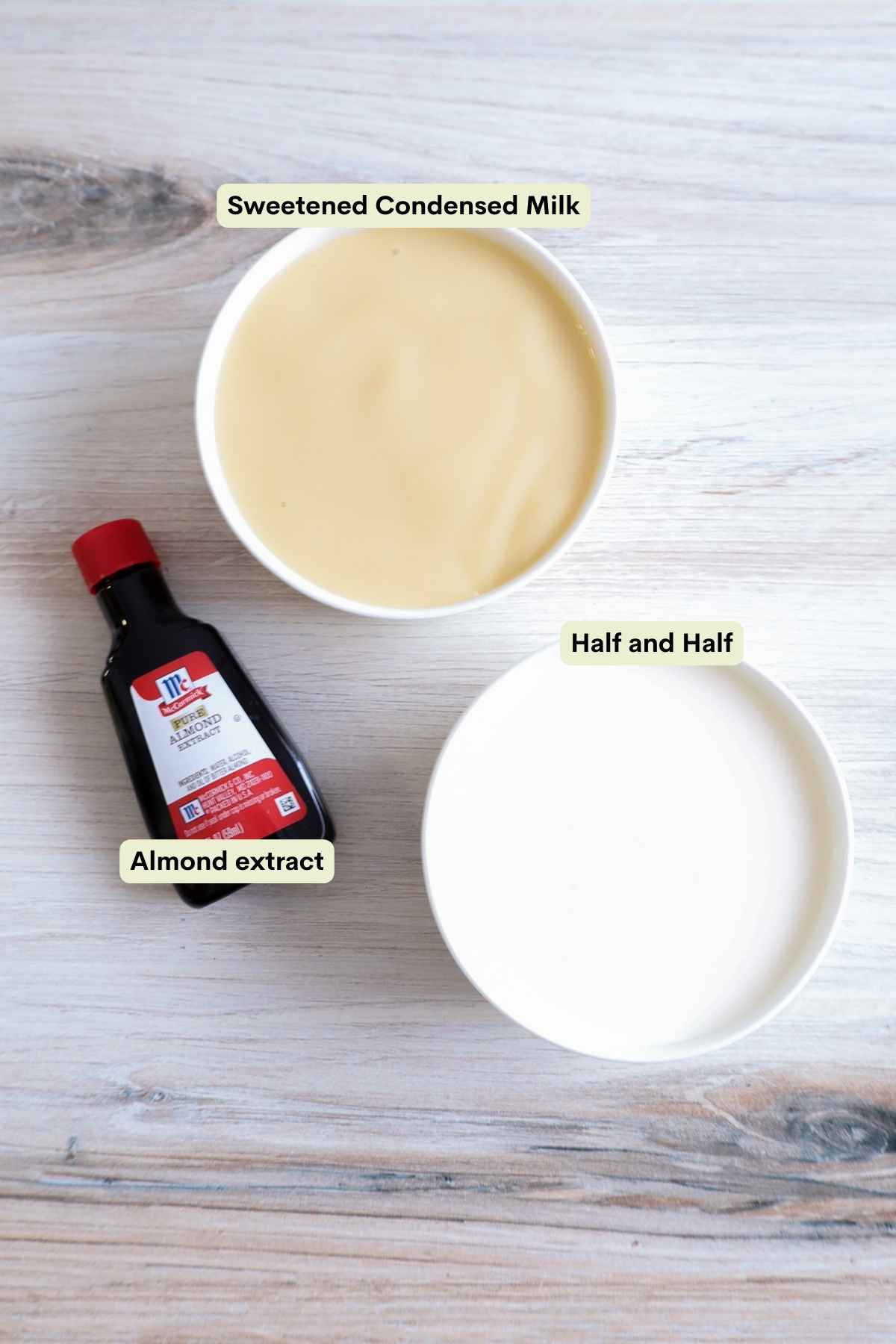 Flat lay of ingredients; a white bowl filled with sweetened condensed milk, a bottle of almond extract, and a white bowl full of half and half.