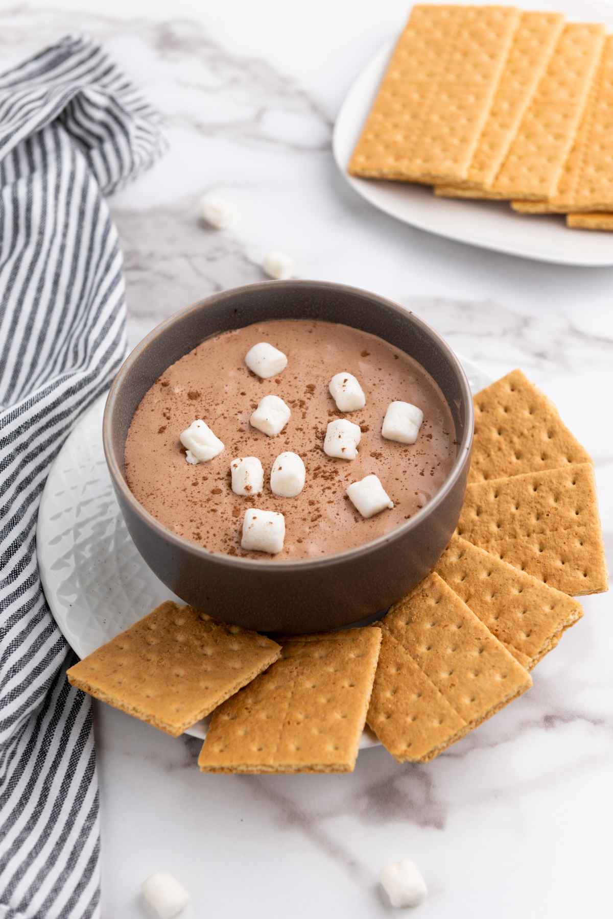 Creamy hot cocoa dip topped with mini marshmallows and cocoa powder in a dark bowl, served with graham crackers on a white plate.