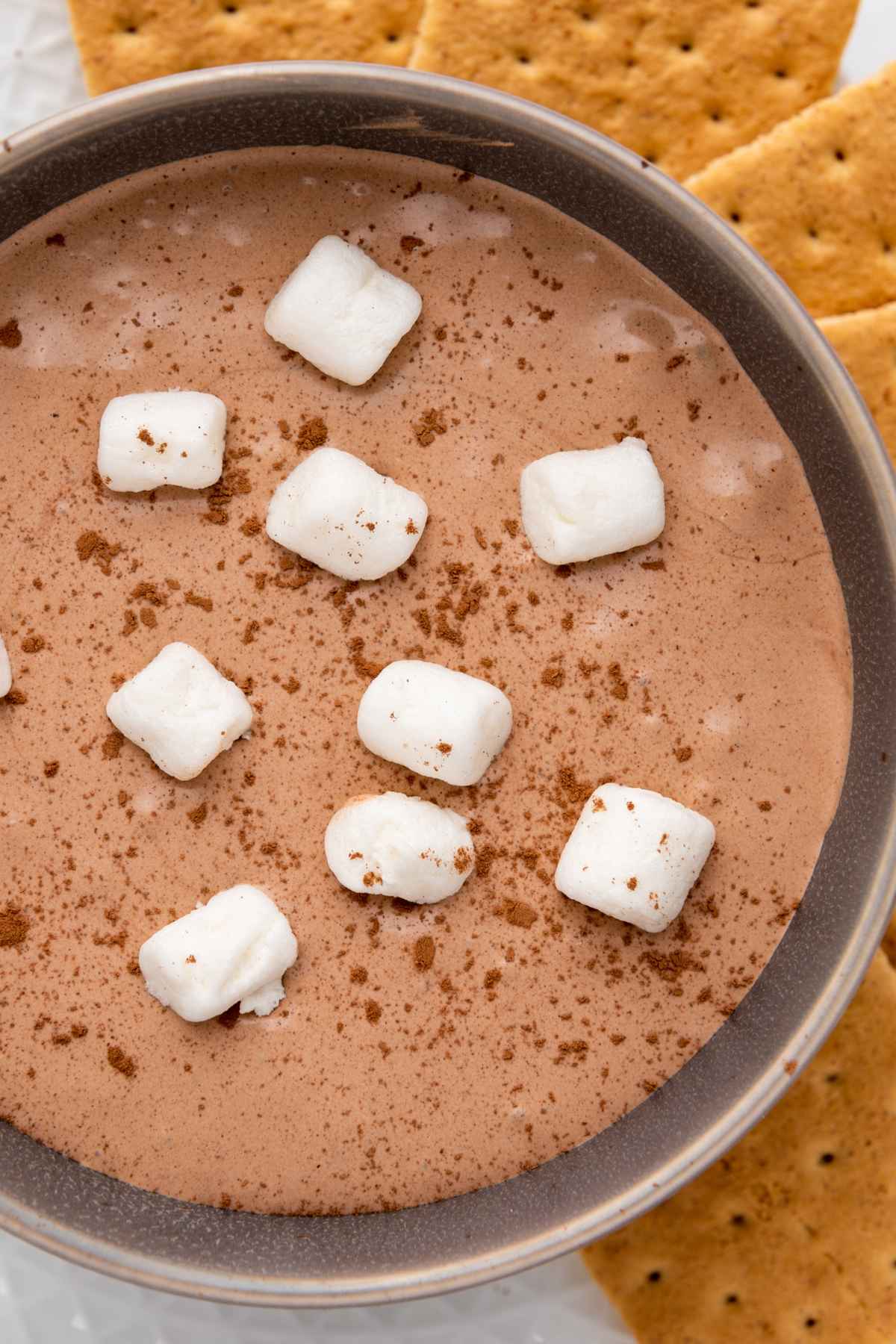 Upclose overhead view of hot cocoa dip topped with mini marshmallows and cocoa powder in a dark bowl, served with graham crackers on a white plate.