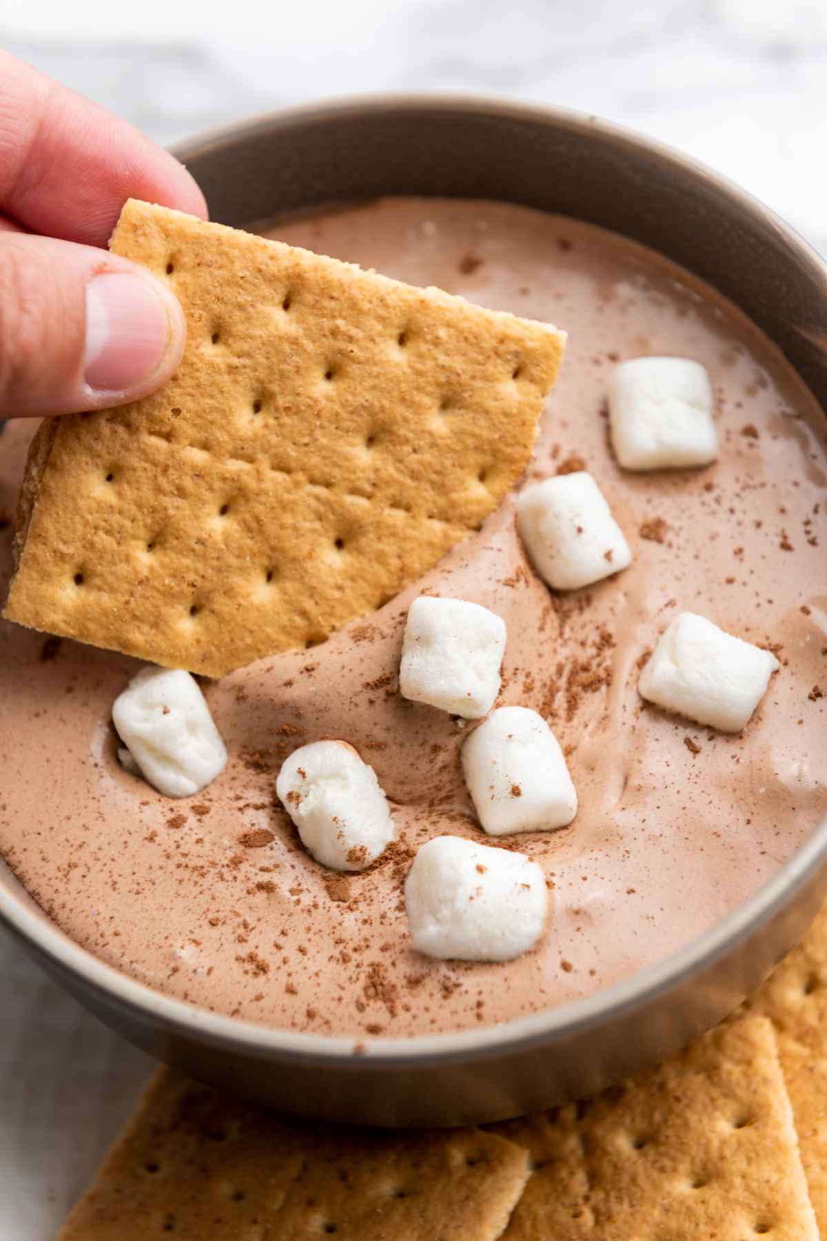 A hand dipping a graham cracker into hot cocoa dip topped with mini marshmallows and cocoa powder in a dark bowl.