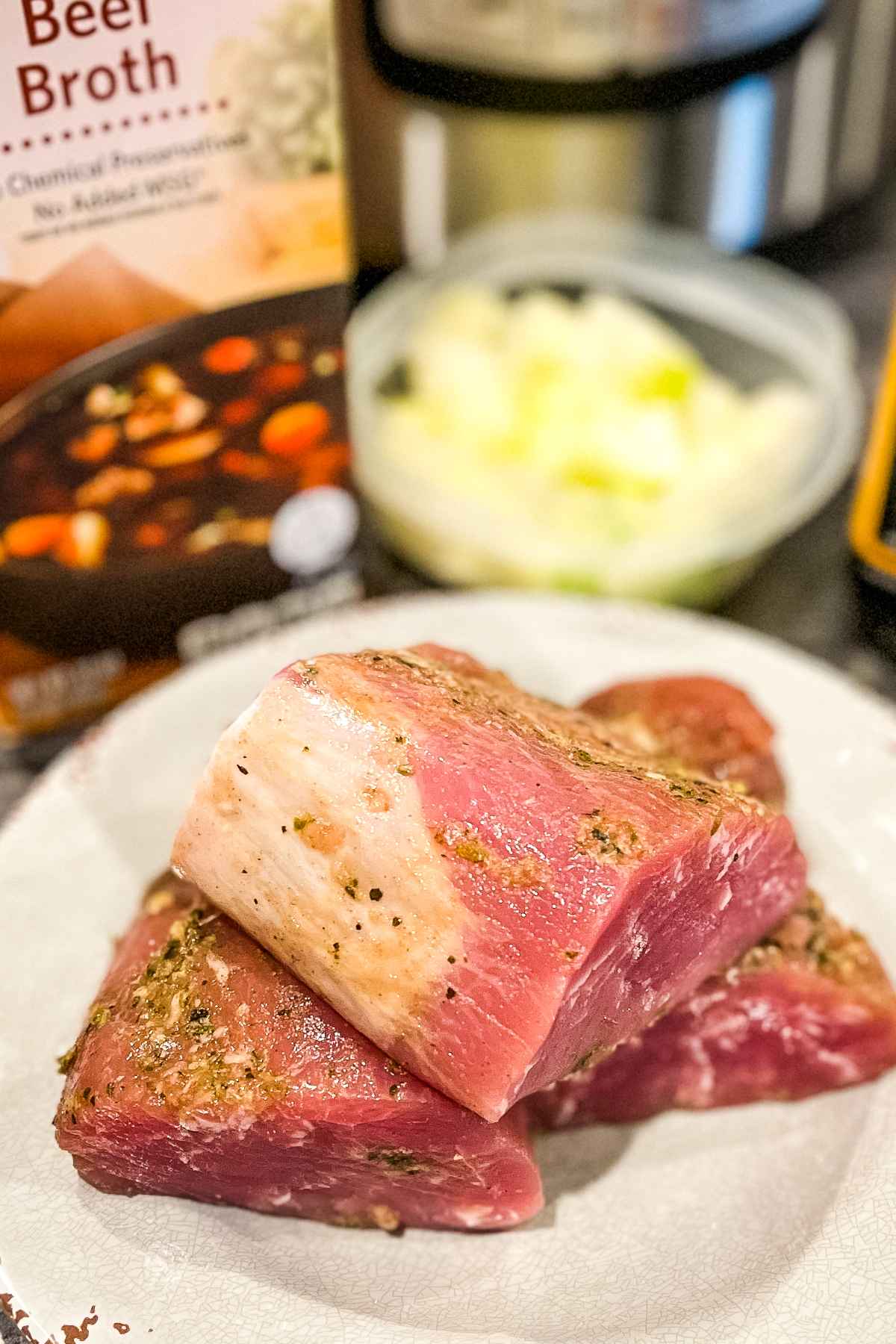 Raw pork tenderloin seasoned and ready to cook, with beef broth and chopped onions in the background.