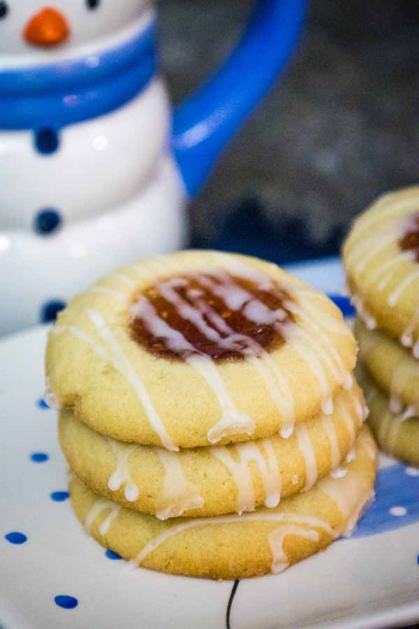 A stack of three raspberry thumbprint cookies on a plate, each topped with jam and a drizzle of icing.