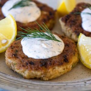 Crispy golden salmon patties topped with a dollop of dill sauce and a sprig of fresh dill, served with lemon wedges.