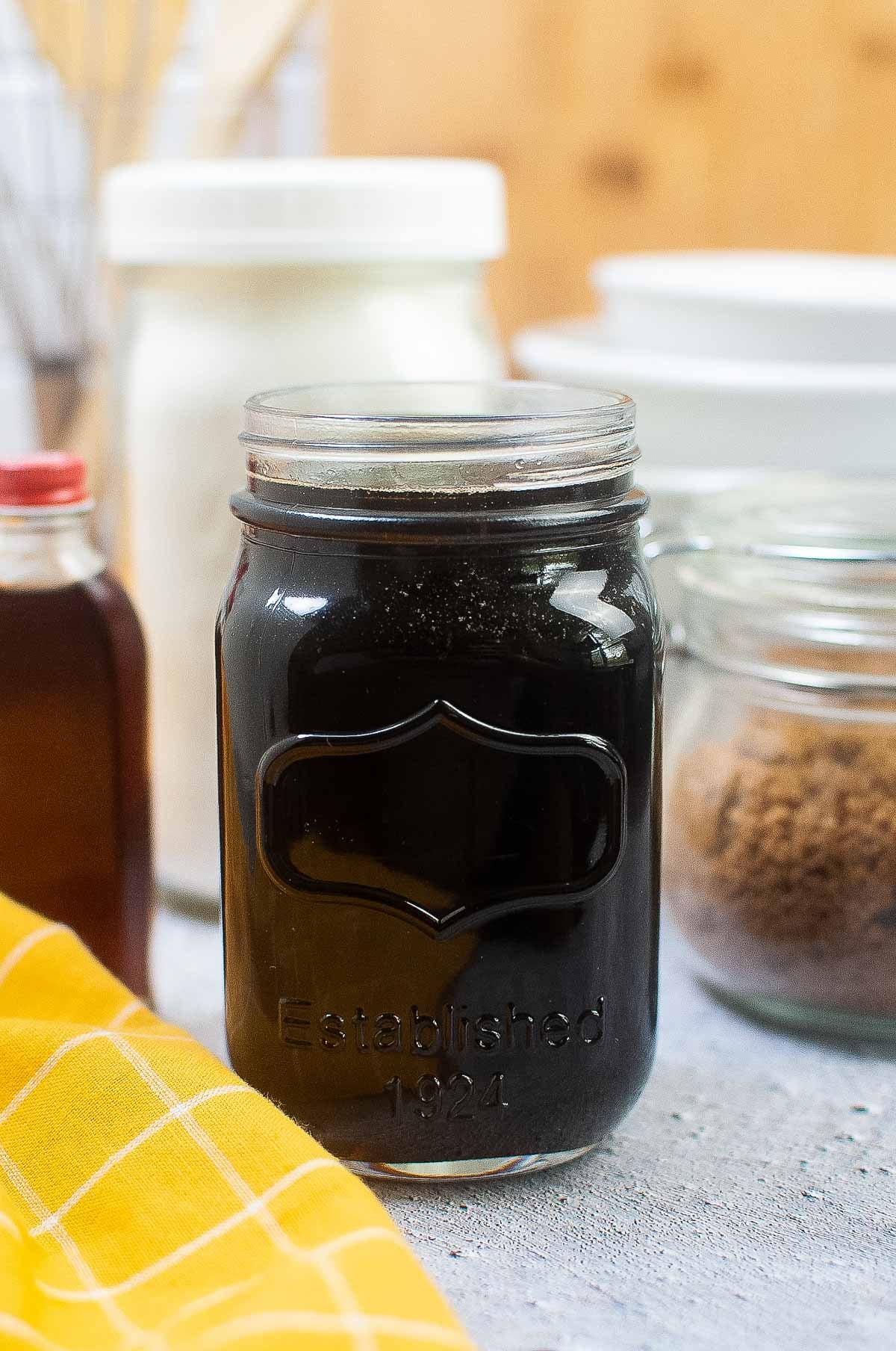 Glass jar filled with homemade pancake syrup, surrounded by baking ingredients and a yellow cloth.