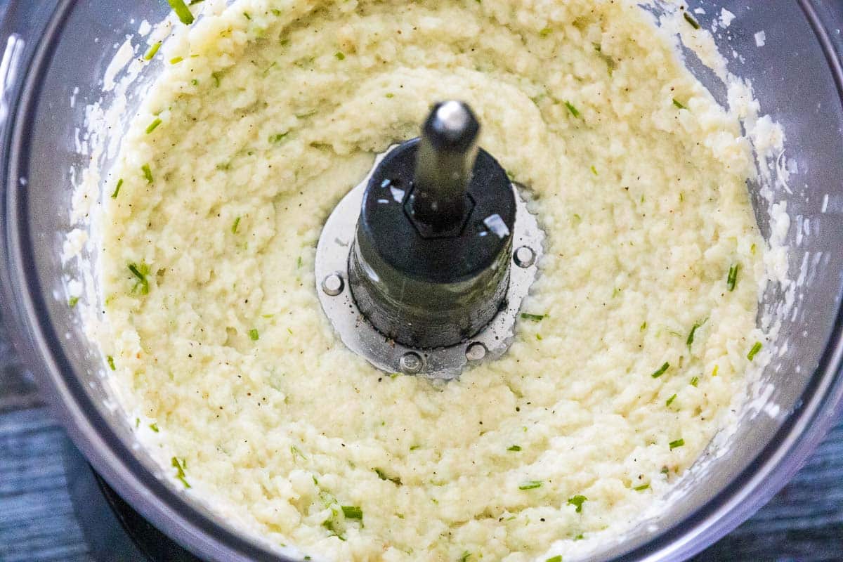 Smooth cauliflower mashed potatoes with green herb flecks in a food processor, ready to be blended.