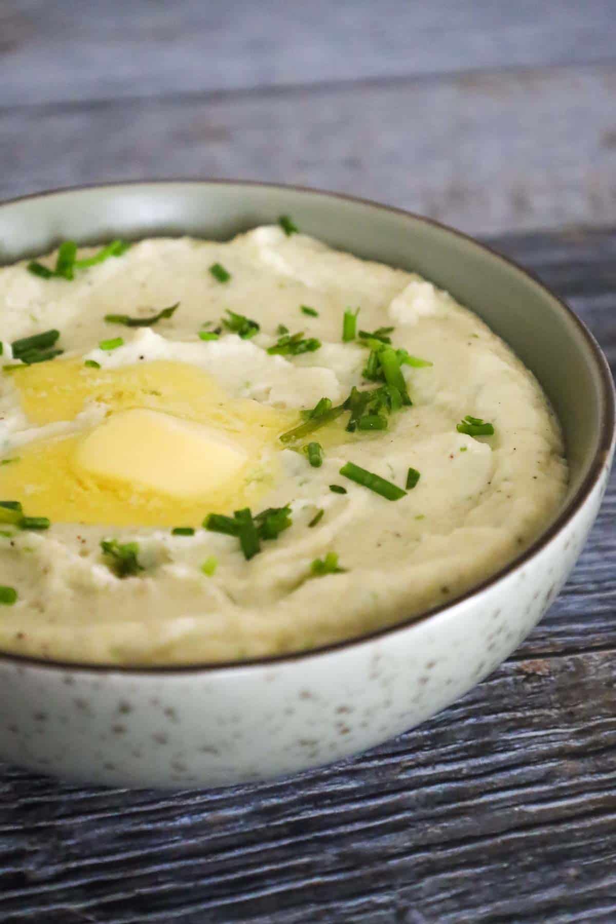A bowl of creamy mashed cauliflower topped with melted butter and garnished with chopped fresh herbs.