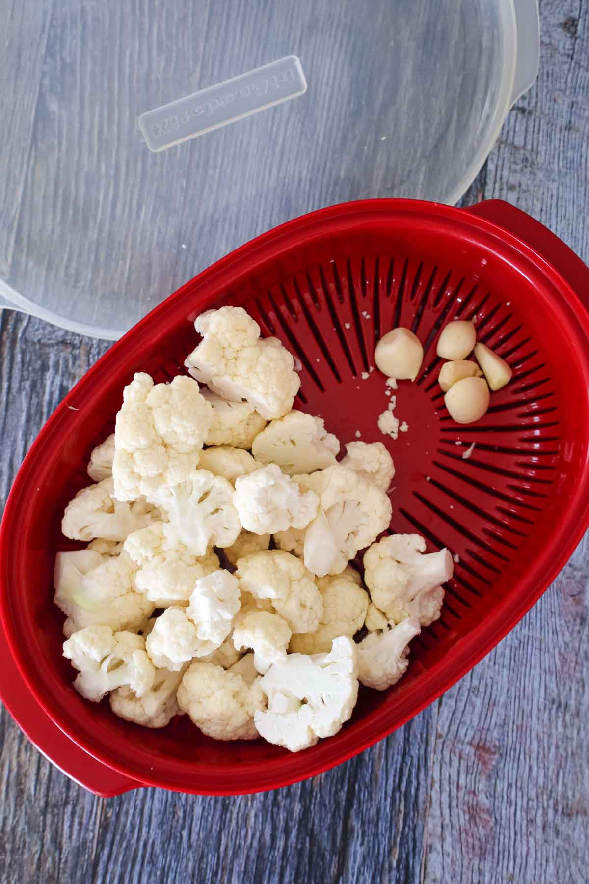 Raw cauliflower florets and garlic cloves in a red microwave steamer basket, prepped for cooking.