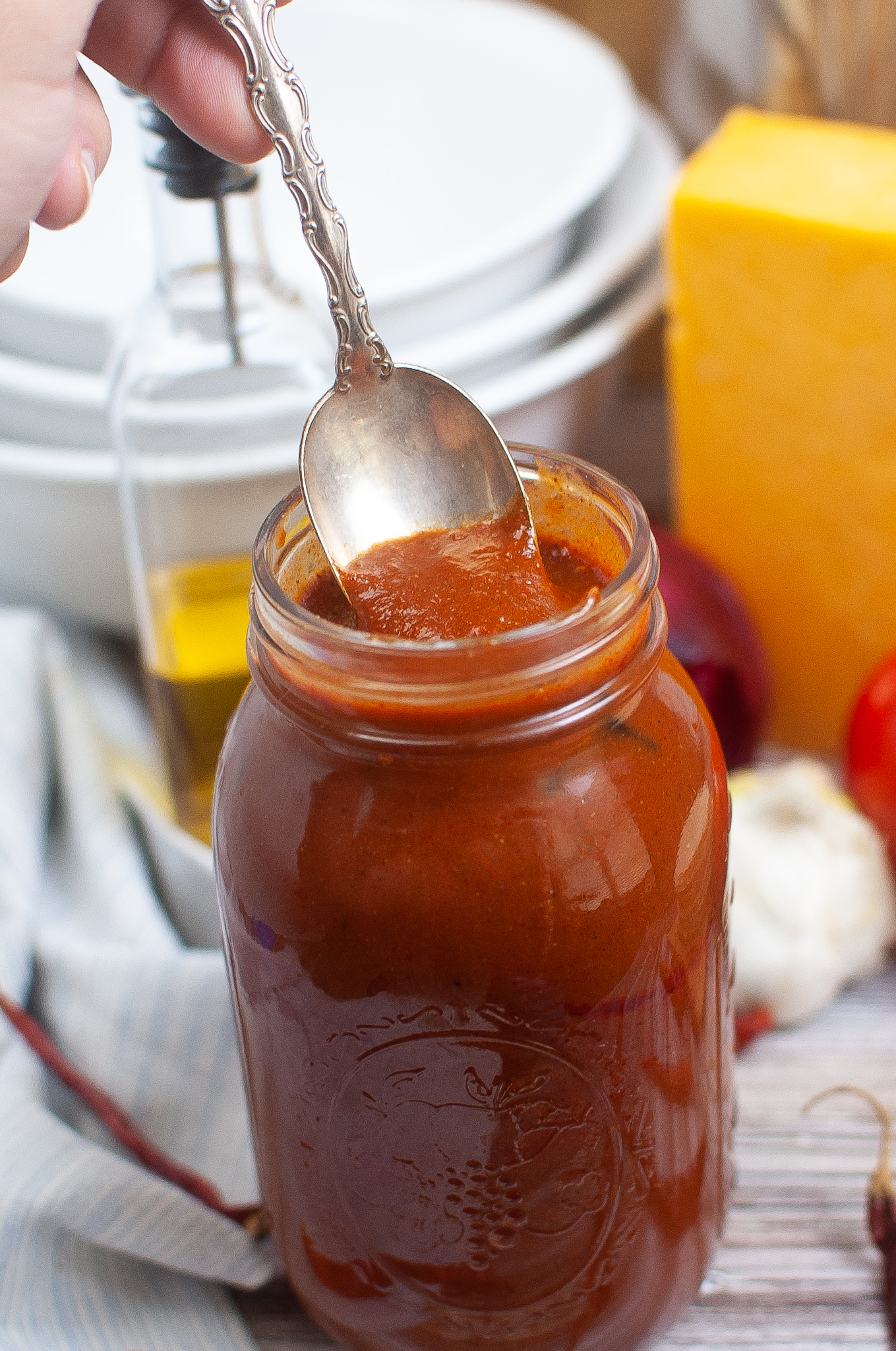 A hand holds a spoon dipping into a glass jar filled with thick red enchilada sauce, with olive oil, cheese, garlic, and tomatoes in the background.