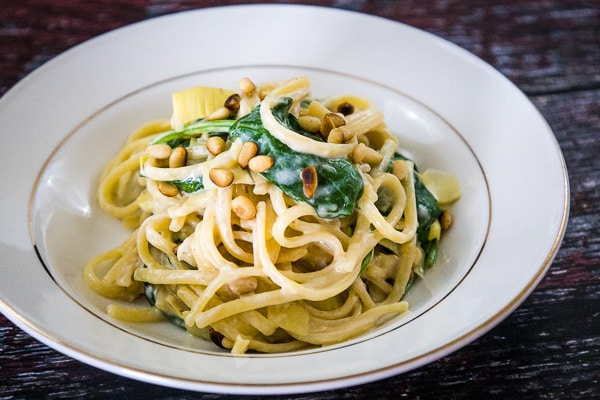 Image of creamy spinach artichoke pasta in a cream gold rimmed bowl garnished with toasted pine nuts.