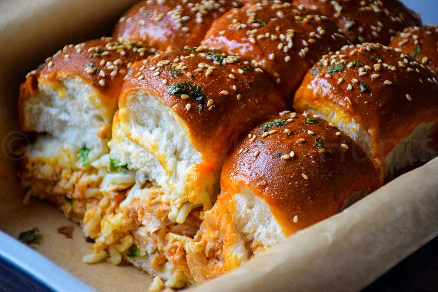 Freshly baked buffalo chicken sliders in a baking dish with golden sesame topped buns, partially pulled apart to reveal the cheesy chicken filling.