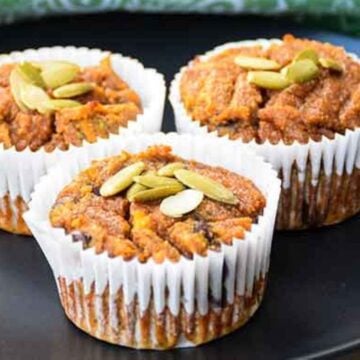 Three paleo pumpkin muffins in white paper liners, topped with pepitas, on a black plate.