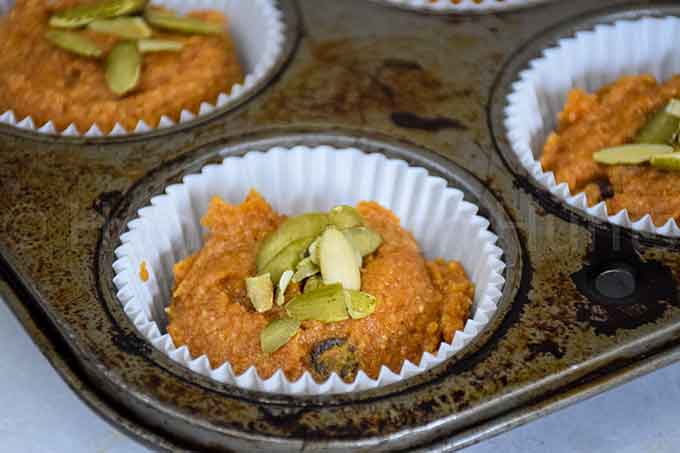 Unbaked muffin batter in paper liners in a muffin tin, topped with raw pepitas.