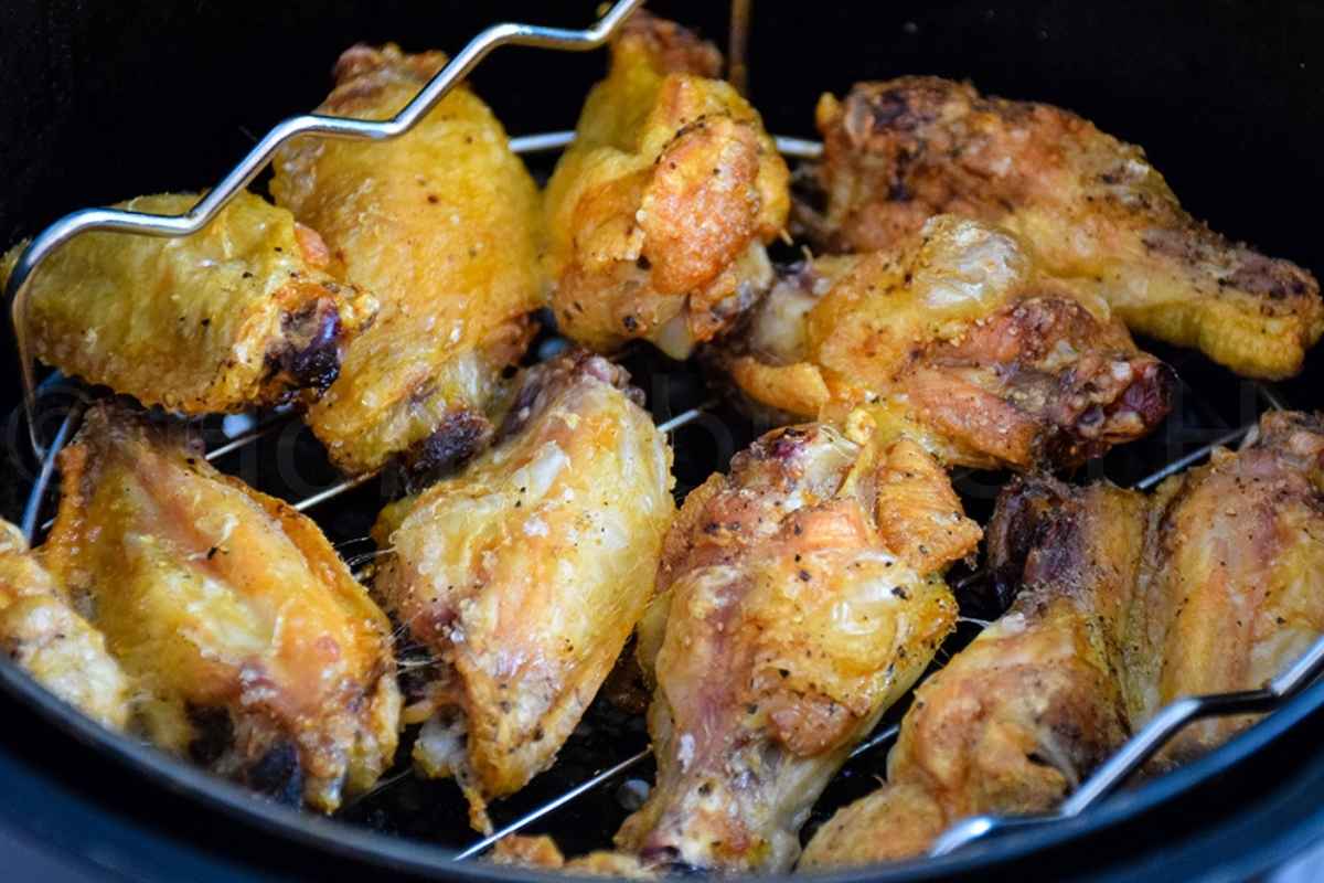 Close up of crispy, golden brown chicken wings cooking in an air fryer basket, seasoned lightly with salt and pepper, with visible grill rack inside the fryer.