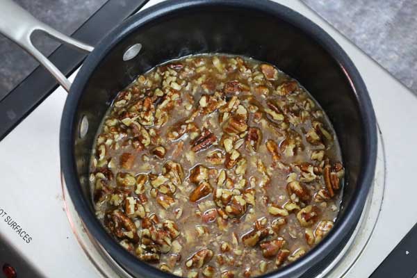 Chopped pecans simmering in a saucepan with a caramel like mixture on a stove top.