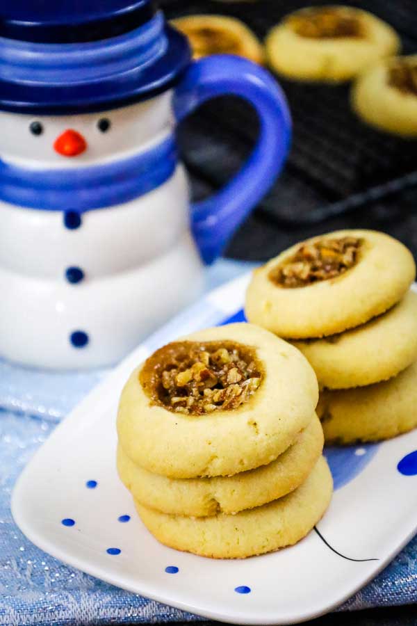 Stack of baked pecan pie thumbprint cookies on a white plate with blue dots, next to a snowman shaped mug.