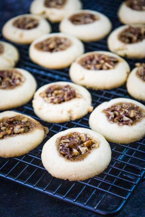 Freshly baked pecan pie thumbprint cookies cooling on a black wire rack, showing golden edges and pecan filling.