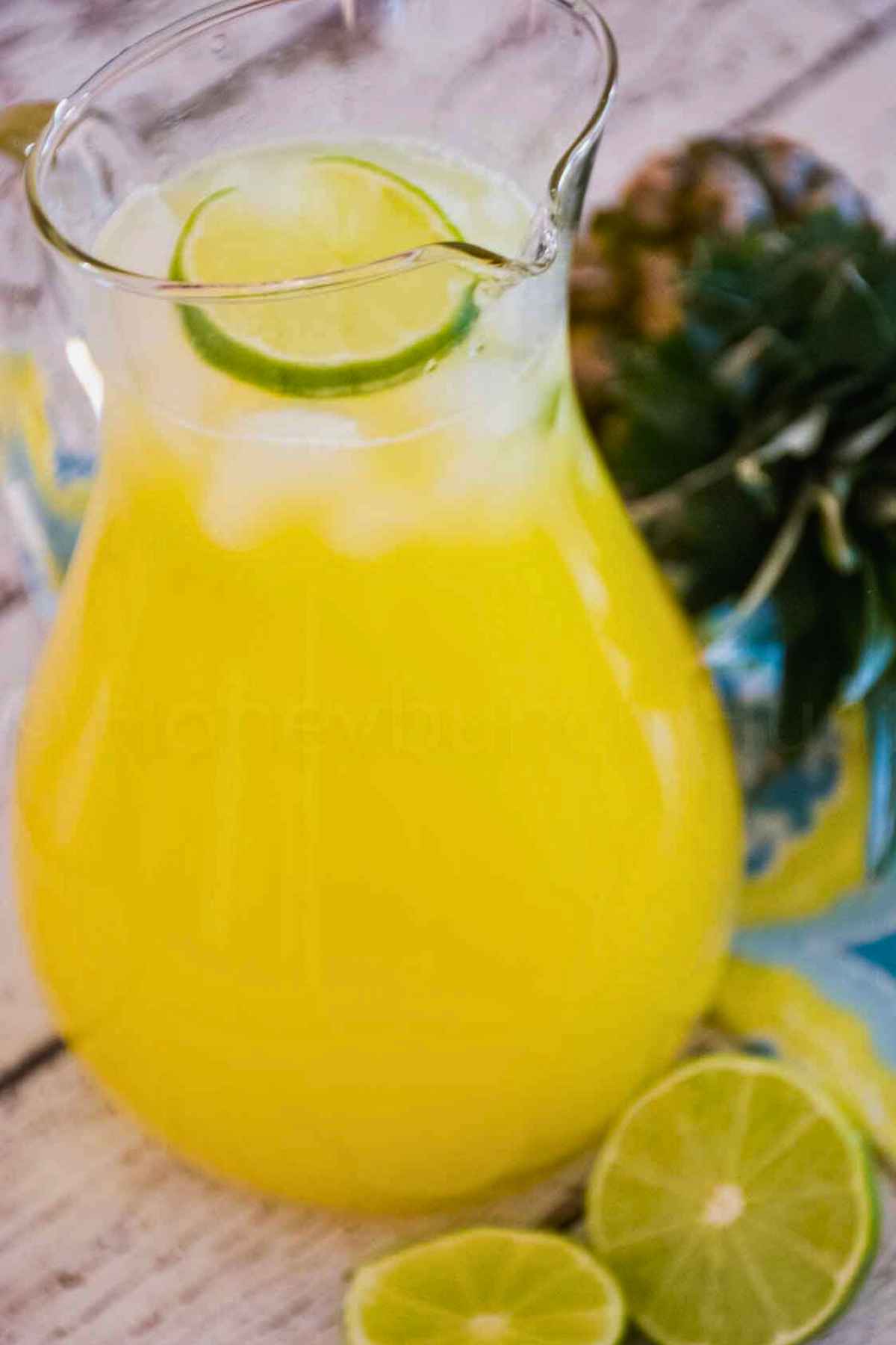 Glass pitcher filled with pineapple agua fresca, garnished with lime slices, with fresh limes nearby.