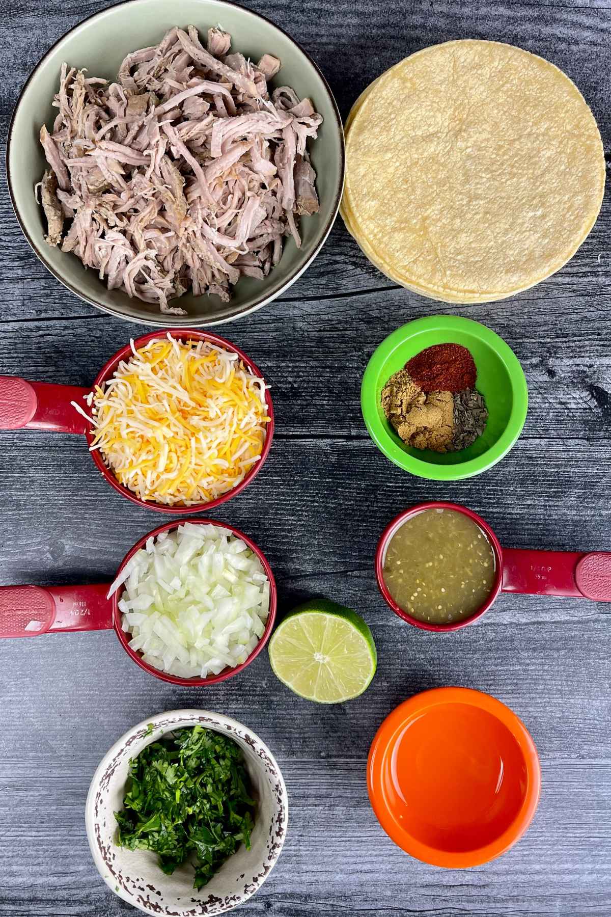 Ingredients for pulled pork taquitos including shredded pork, tortillas, cheese, onion, salsa, spices, lime, and cilantro