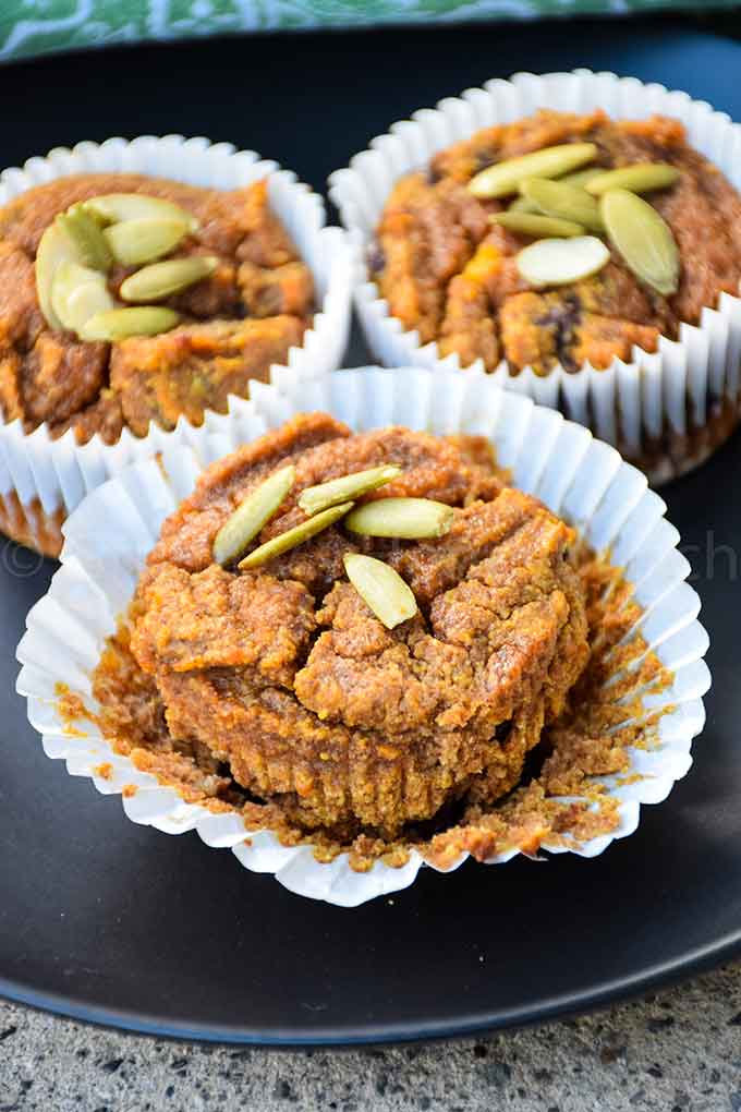 Three paleo pumpkin muffins in white paper liners, topped with pepitas, on a black plate.